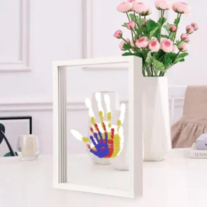 Handprint art with overlapping colorful prints displayed in a white frame on a bright, modern tabletop next to a vase of flowers.
