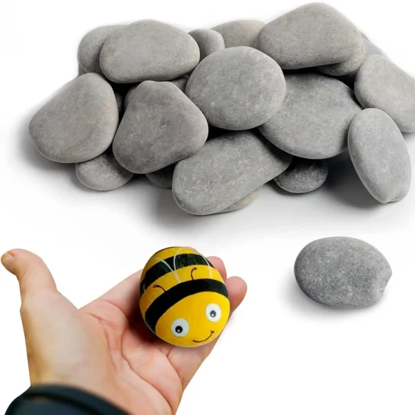 A hand holding a bee-painted stone, with a pile of smooth grey painting rocks in the background.